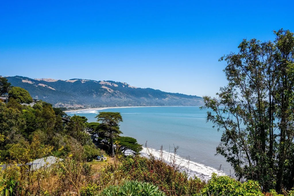 Coastal view of a beach, ocean, hills, and trees on a bright, clear day.