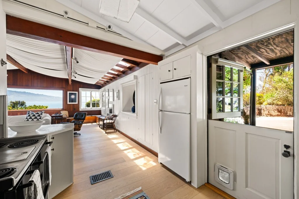 Open kitchen with white cabinets and refrigerator, view into living room with wooden wall, windows, and outdoor scenery beyond.
