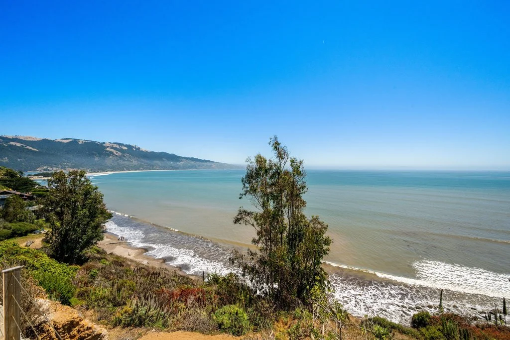 A view of a beach with waves hitting the shore, green trees and bushes in the foreground, and a distant coastline with hills, under a clear blue sky.
