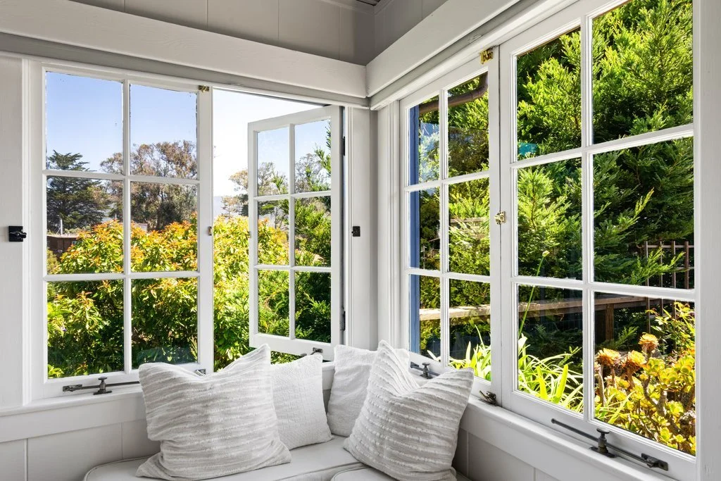 Sunlit interior with white cushioned seating and view of lush green garden through multiple large windows.