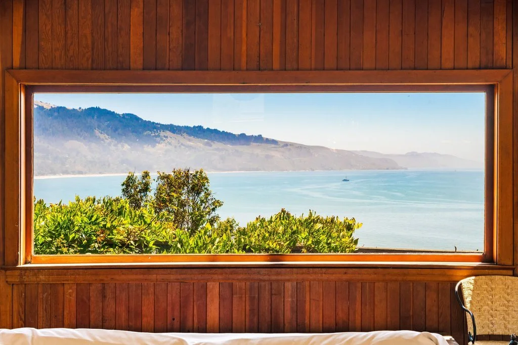 View of the ocean and distant hills through a large rectangular window with wooden frame, inside a room with wooden walls and a chair.
