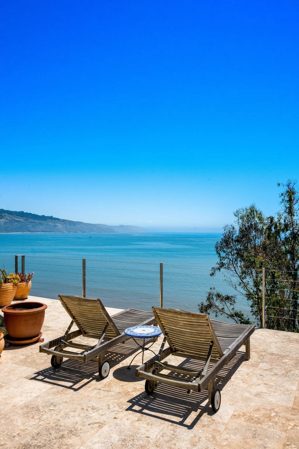 Two wooden lounge chairs on a stone patio overlooking a calm ocean with a distant shoreline and clear blue sky.