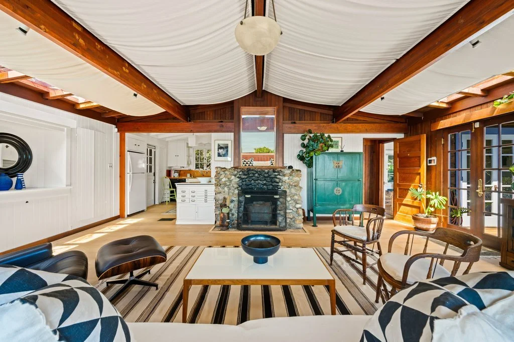 Living room with a stone fireplace, wooden beams, and large windows, with furniture including a white sofa, wooden chairs, and a black leather chair.