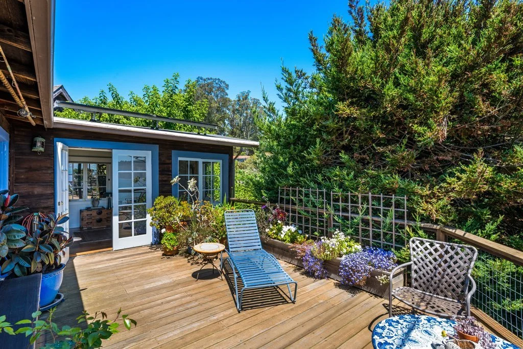 A wooden deck garden patio with a lounge chair, a metal chair, a small side table, and potted plants and flowers, adjacent to a house with open glass doors, surrounded by green trees and a clear blue sky.