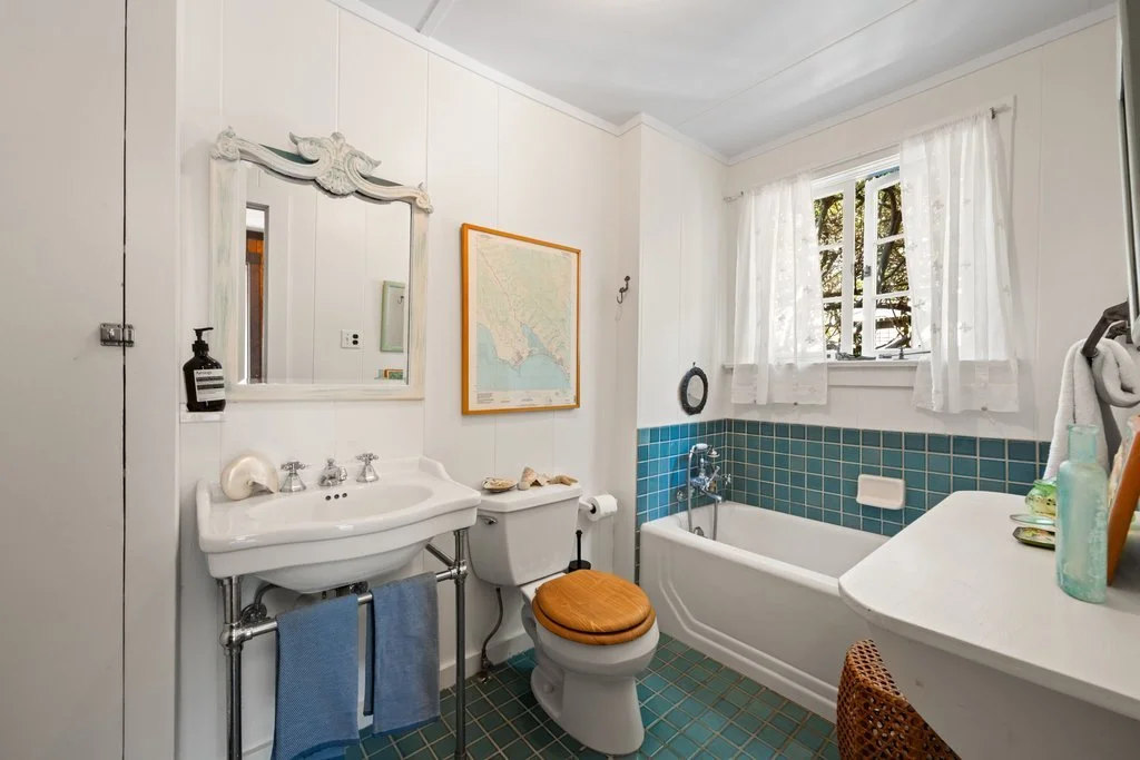 Bathroom with white walls, a window with white curtains, a bathtub with turquoise tiles, a white sink with a mirror, a toilet with a wooden seat, and a small white counter with decorative bottles.