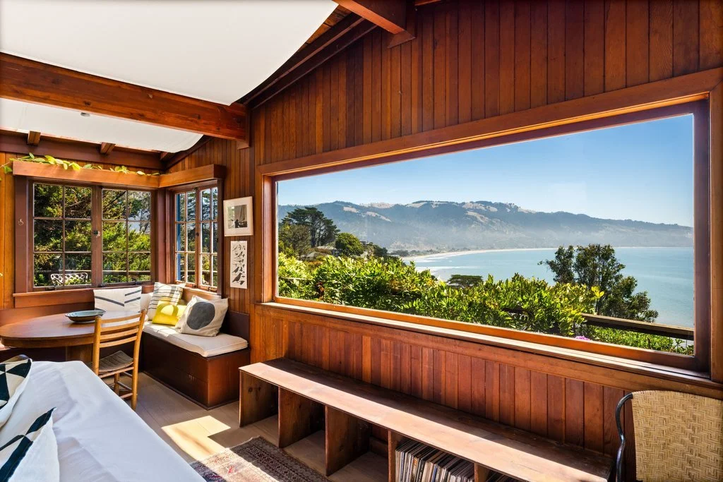 Cozy room with wooden walls and large windows showcasing a view of mountains, trees, and ocean.
