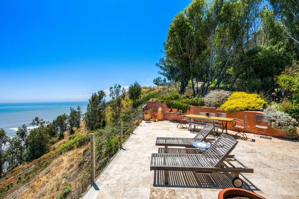 Outdoor patio on a hillside overlooking the ocean with lounge chairs, a table, and potted plants, surrounded by trees and greenery.
