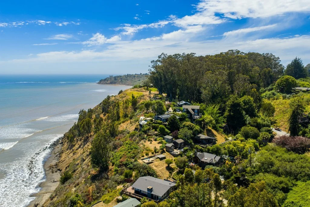 Aerial view of a coastal hillside with houses, surrounded by lush green trees, overlooking the Pacific Ocean under a partly cloudy sky.
