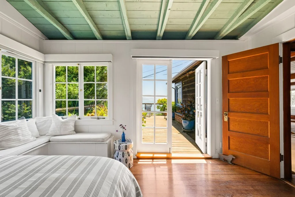 Interior of a bright bedroom with white walls, large windows, and a door that opens to a deck with outdoor furniture and ocean view.