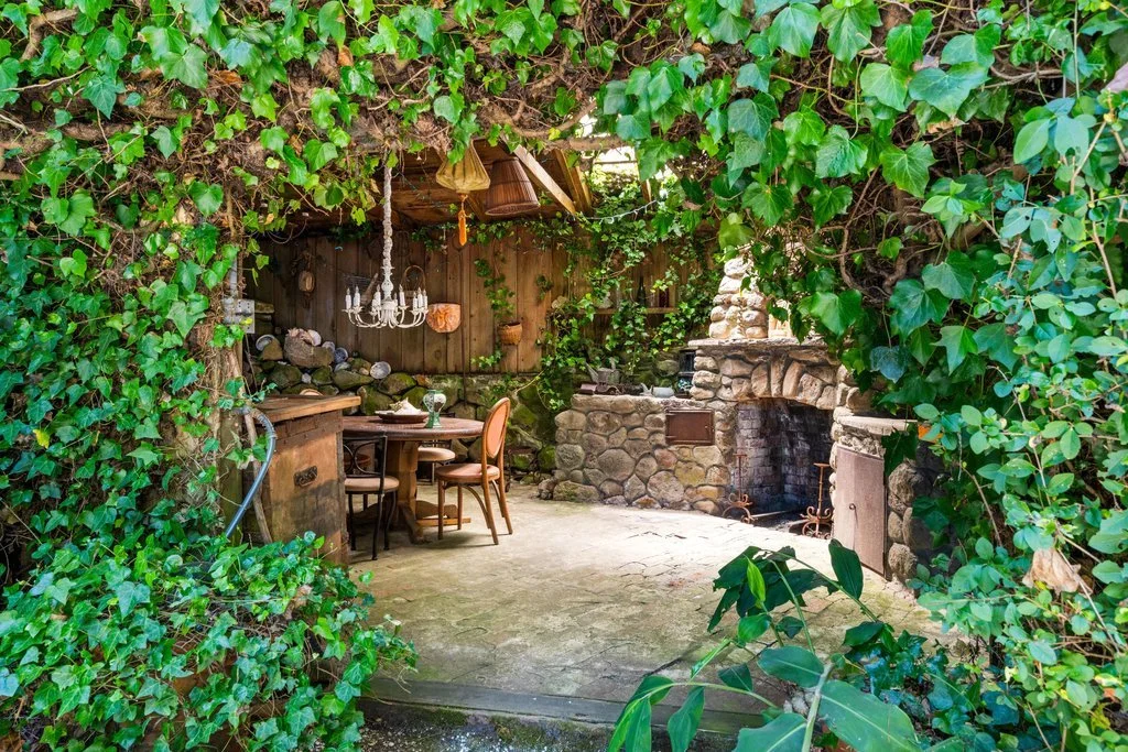 A cozy outdoor space surrounded by lush green ivy, featuring a stone fireplace, a round wooden dining table with four chairs, and a chandelier hanging from above, creating a rustic, inviting atmosphere.