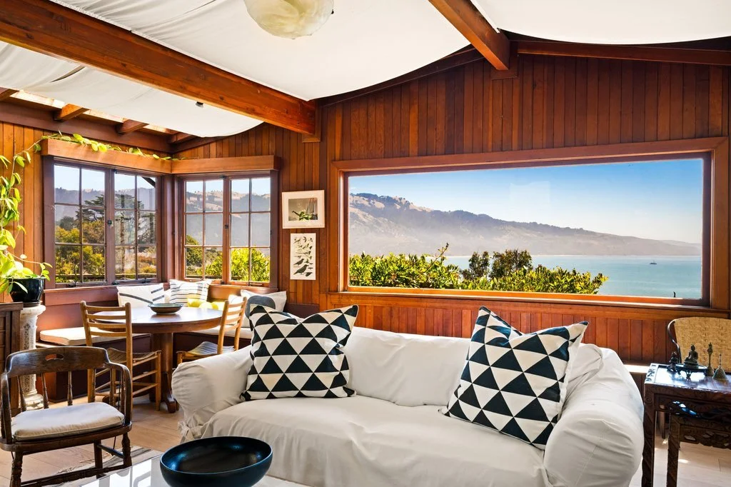 A cozy living room with wood-paneled walls and large windows showcasing a view of a mountain, trees, and ocean. The room has a white sofa with patterned pillows, a round dining table with chairs, and potted plants.