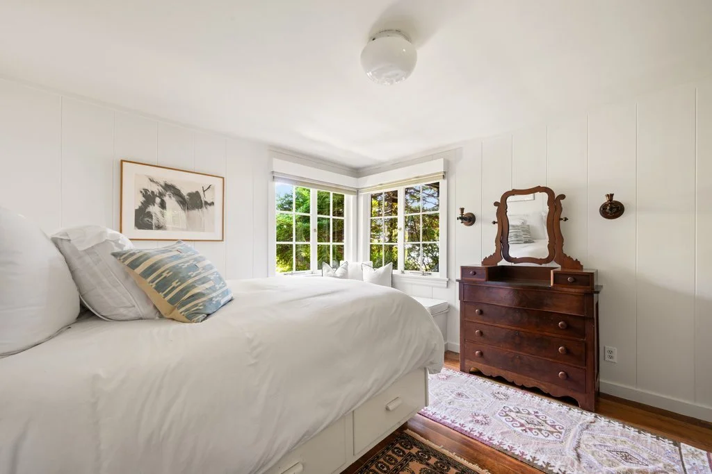 A cozy bedroom with a large bed, white bedding, and decorative pillows. There is a wooden dresser with a mirror and wall-mounted lamps, a window with a view of trees, and a colorful area rug on the wooden floor.