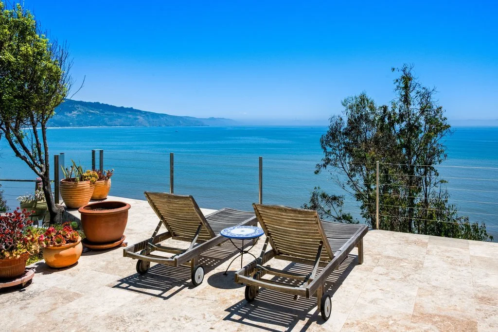Two wooden lounge chairs with wheels on a terrace overlooking the ocean, with potted plants and trees nearby, and a clear blue sky.