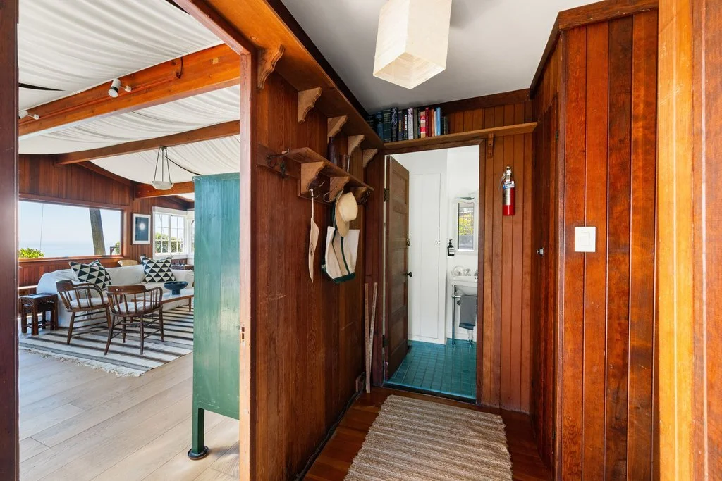 Wood-paneled hallway with view into a living room with large windows, dining table, and chairs, and a view of the outdoor scenery. A small bathroom with green tiled flooring is visible through the open door. Hooks on the wall hold hats and bags, with