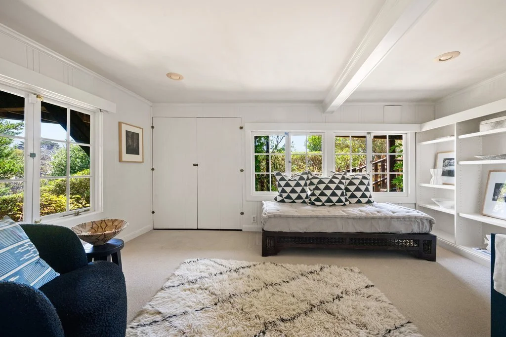 Bright bedroom with white walls and large windows showing greenery outside. Contains a bed with decorative pillows, a black armchair, a woven basket, a white bookshelf with framed photos, and a shaggy cream rug.