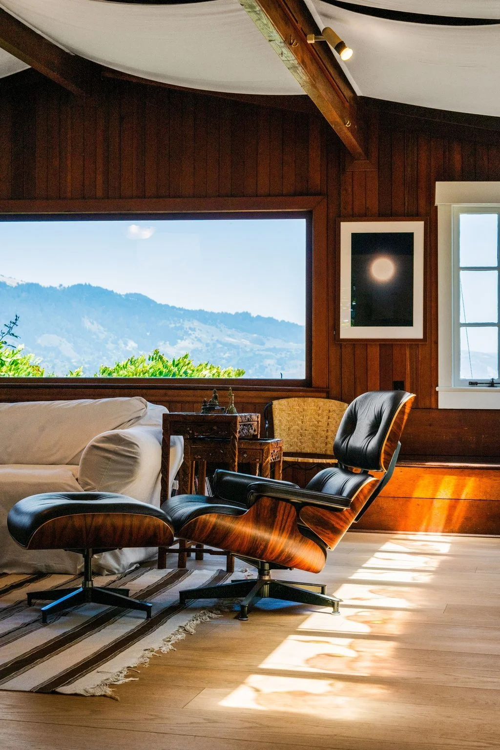 Interior of a room with wood-paneled walls, featuring a large window showing a mountain landscape, a black leather chair, a white couch, and a framed moon photograph on the wall.