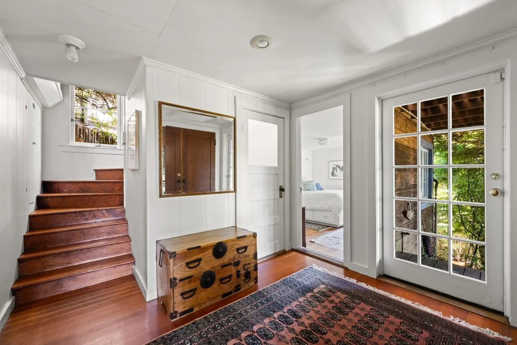 A cozy interior with white walls, wooden stairs leading up, a mirror above a chest, a glass-paned door to the outside, and a view into a bedroom with a bed and artwork.