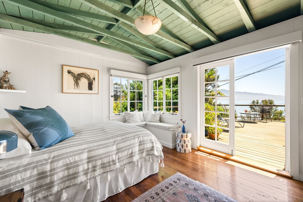 A cozy bedroom with white paneled walls, a bed with striped bedding and blue pillows, a window seat with cushions, and a large glass door opening to a deck with outdoor seating overlooking water and trees.