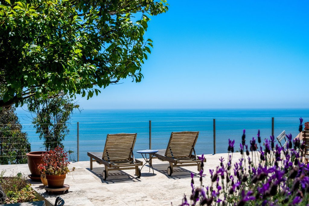 Two wooden lounge chairs on a tiled patio with a small side table between them, overlooking a blue ocean with a clear sky, surrounded by potted plants and purple flowers, and a green tree on the left.