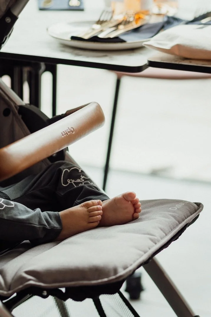 Close-up of a child's bare feet resting on a cushioned high chair in a restaurant, with a table set with plates, utensils, and napkins in the background.