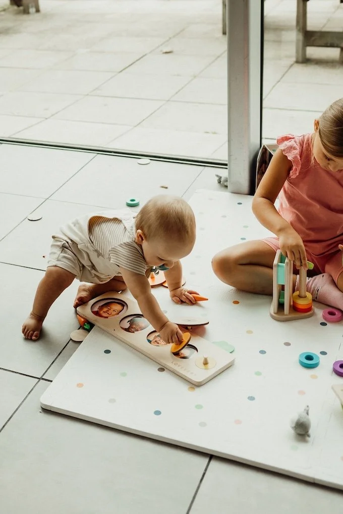 A baby and a young child play with wooden toys on a white mat with pastel dots near a glass door.