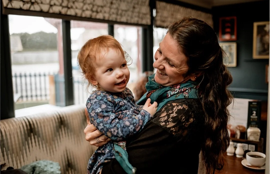 A woman with long curly dark hair laughing and holding a young girl with red hair and a blue floral dress indoors near large windows.