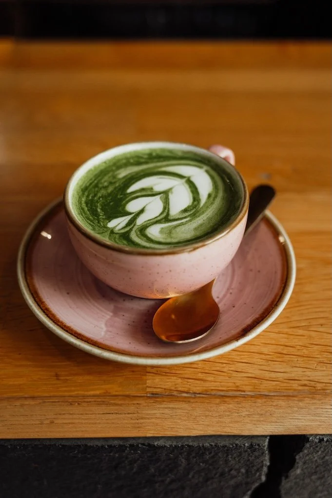 A pink ceramic cup filled with green matcha latte with white latte art on top, placed on a matching pink saucer with a small spoon on a wooden table.