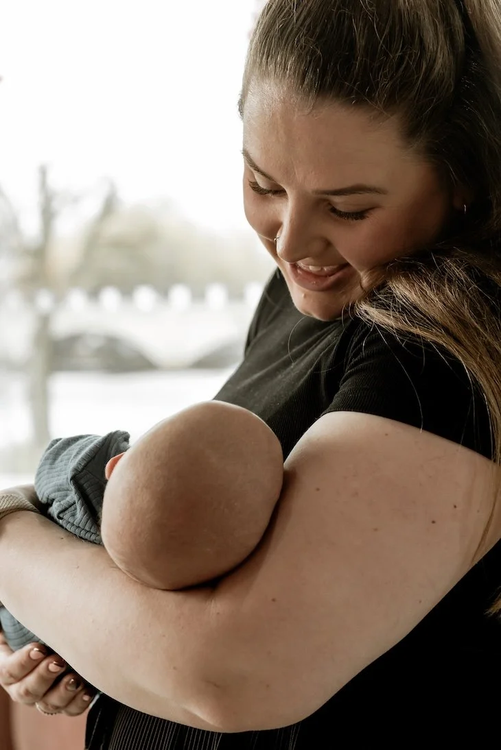A woman with long brown hair smiles while holding a baby in her arms indoors near a window.
