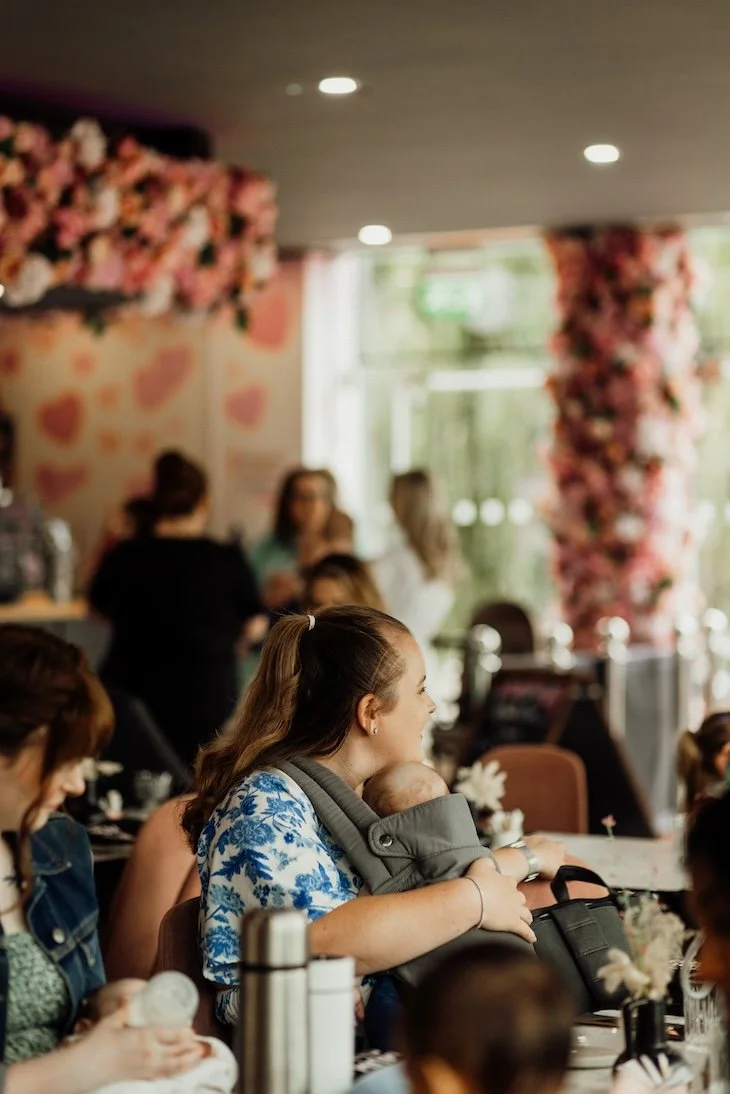 A woman holding a baby sits in a busy cafe decorated with pink flowers; other people chat and dine around her.