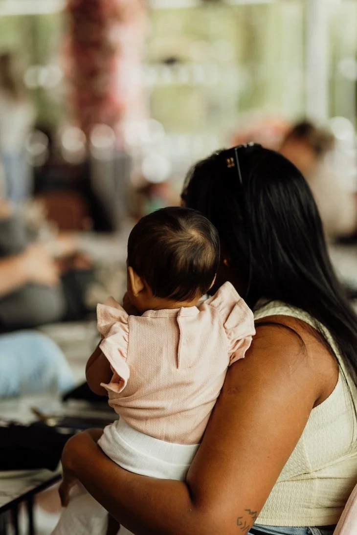 Woman holding a baby at a social gathering or event in an indoor setting with large windows and blurred background.