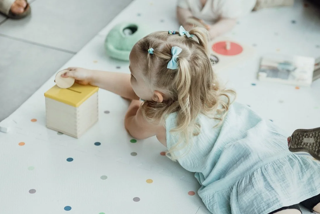 A young girl with blonde hair decorated with blue bows is reaching out to touch a yellow and white block on a table with colorful dots, possibly participating in a playing or learning activity.