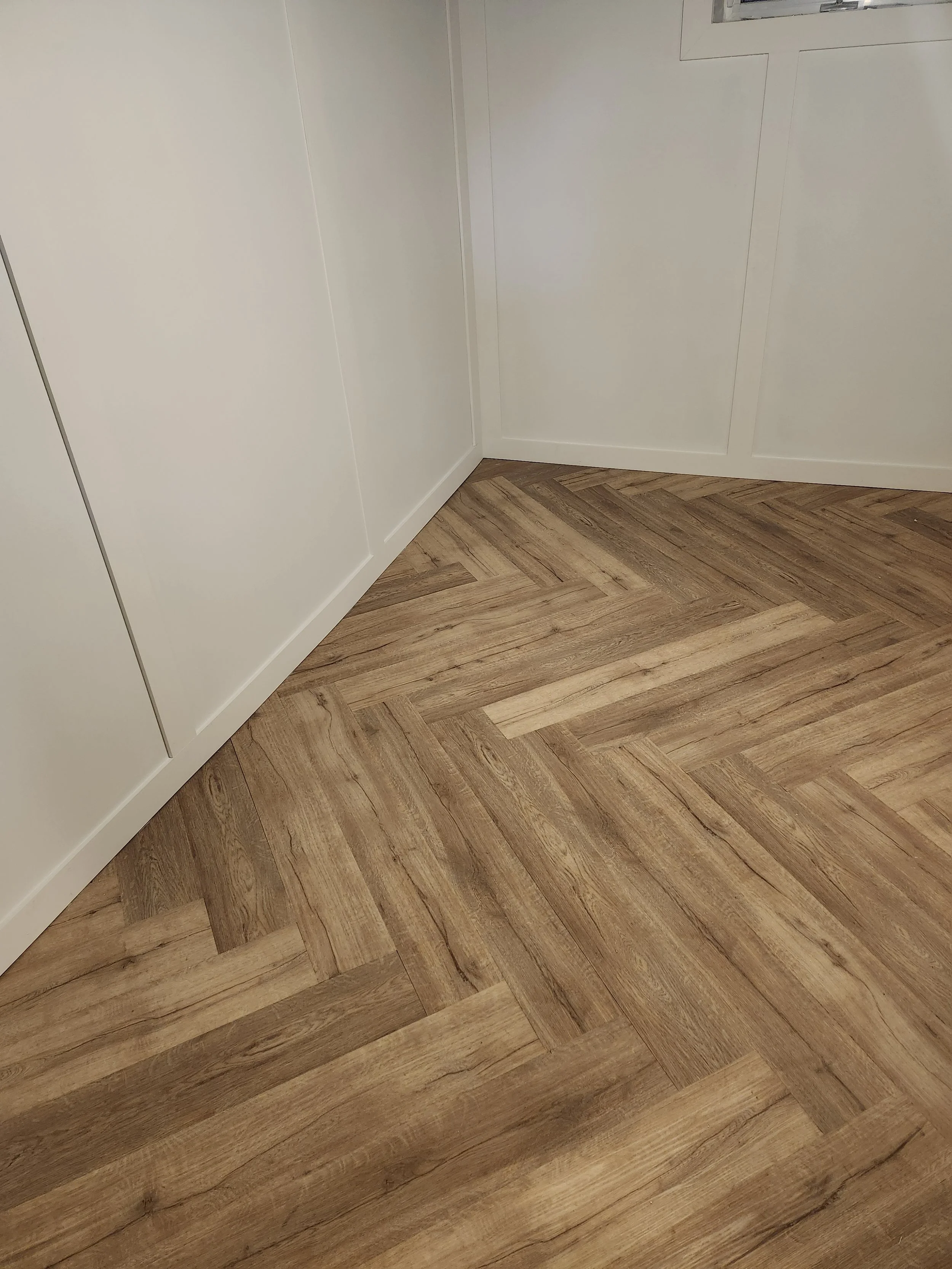 Corner of a room with white walls, baseboards, and wooden chevron-patterned floor.