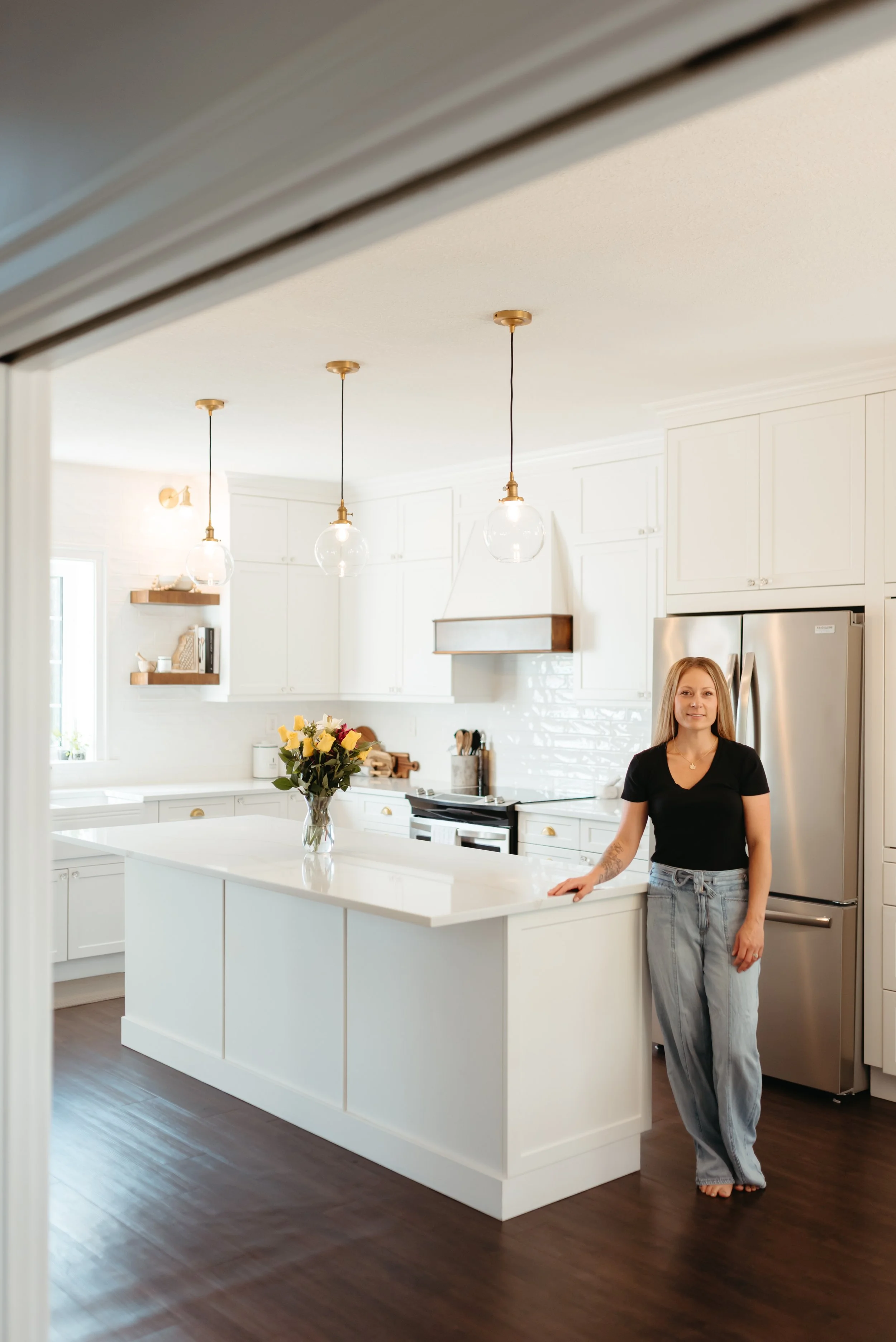 A woman with blonde hair, wearing a black t-shirt and loose-fitting jeans, stands barefoot in a bright, modern kitchen with white cabinets, a white island with a bouquet of flowers, and stainless steel refrigerator, looking at the camera.