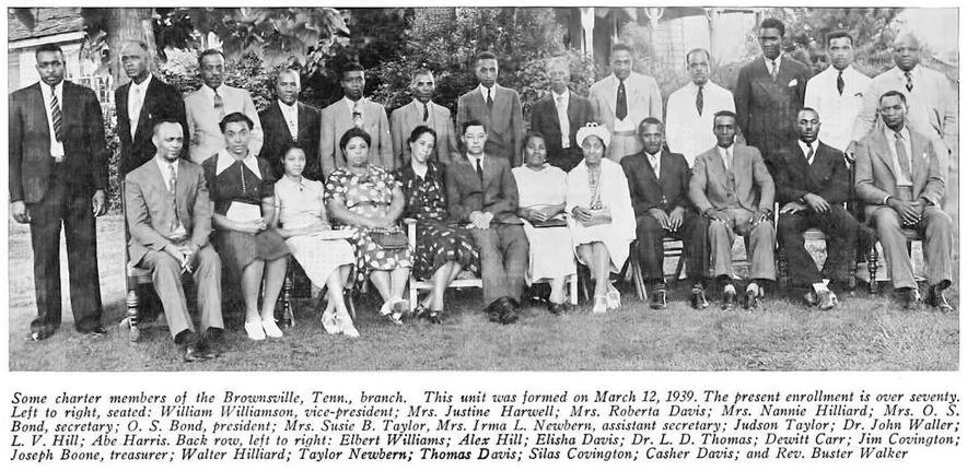 Black and white photo of a large group of men and women, some seated and some standing outdoors on a grassy area, with trees in the background, taken in 1939 at a Brownsville Tennessee branch.