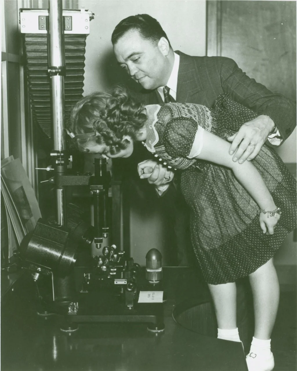 A man in a suit and tie holds a young girl with curly hair as she looks into a microscope on a laboratory table.