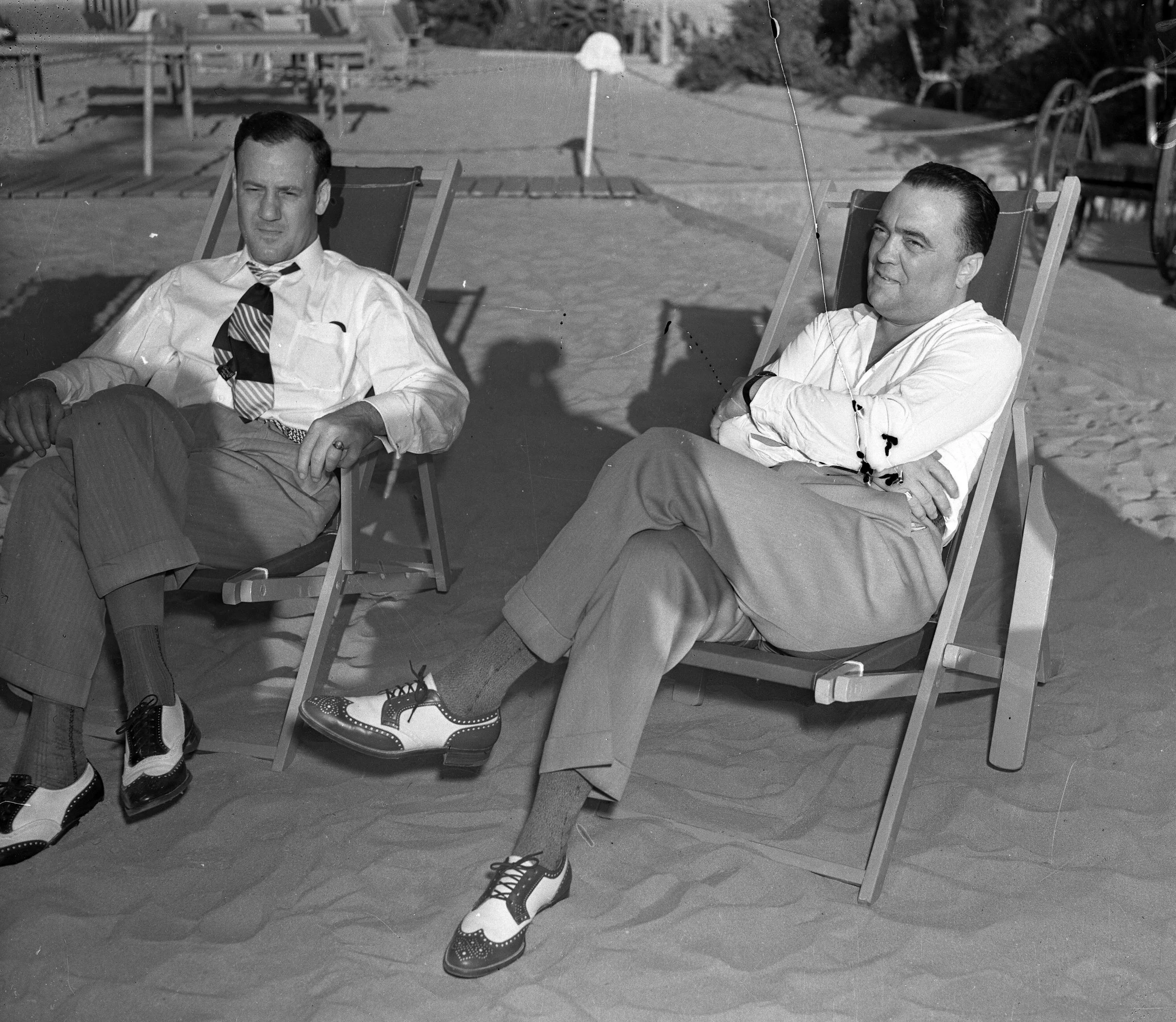 Two men are sitting on lounge chairs outdoors near a beach. Both men are dressed in formal or semi-formal attire, with shirts and trousers. Their shoes are off, placed on the ground in front of them. The man on the left is winking or squinting and has a patterned tie, while the man on the right has his arms crossed and is looking at the camera with a slight smile. In the background, there are umbrellas, trees, and some bicycles.