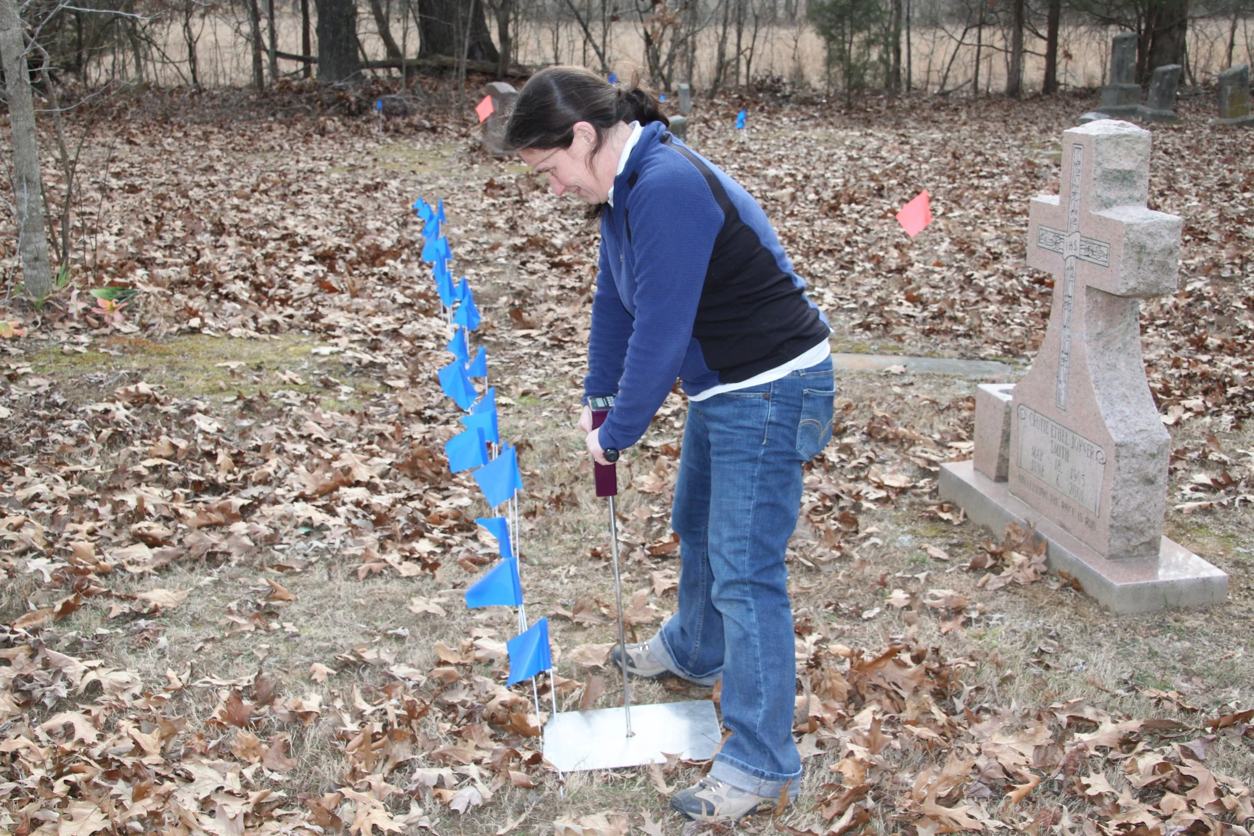 Woman in a blue jacket bending downward at a gravesite with multiple small American flags, in a wooded area with fallen leaves and upright headstones nearby.