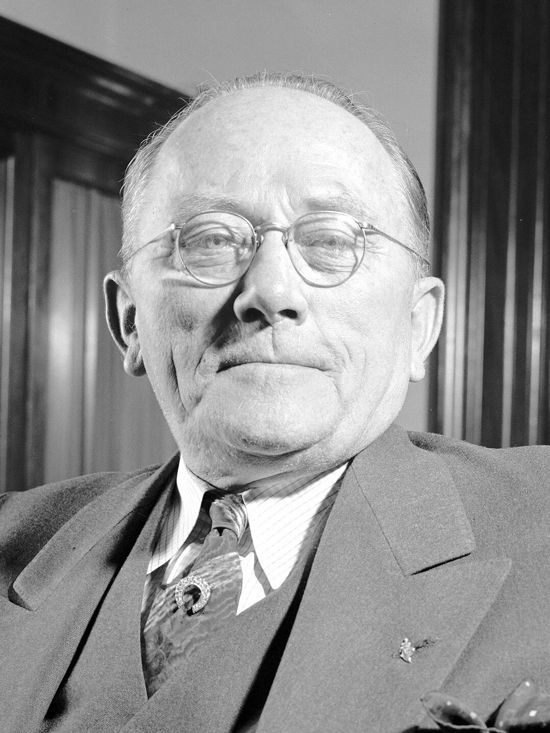 A black-and-white photograph of an elderly man wearing glasses and a suit, with a wooden-paneled background.