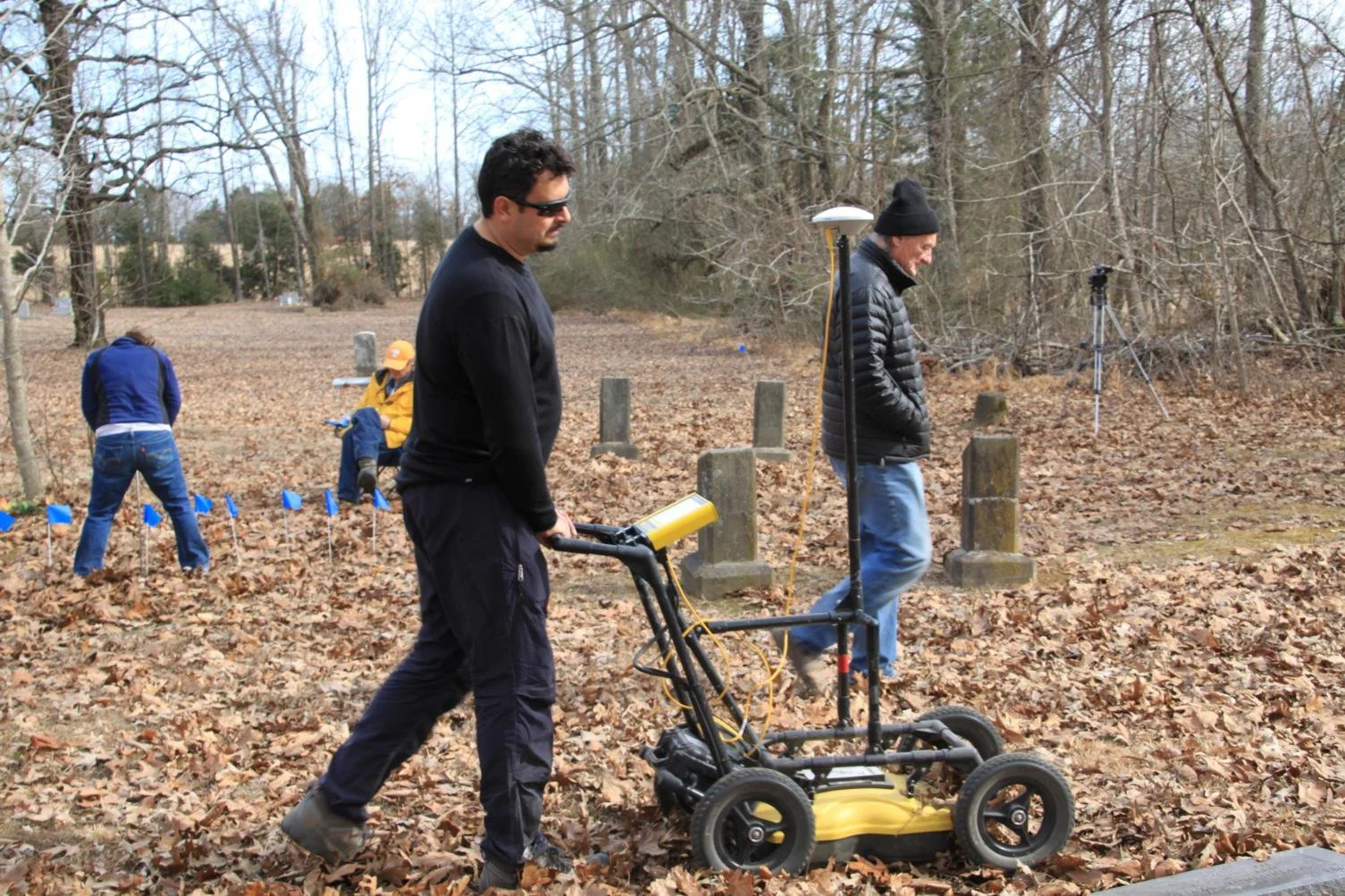Two men working outdoors in a wooded area, one operating a ground-penetrating radar device, others working nearby with equipment, fallen leaves on the ground, leafless trees in the background.