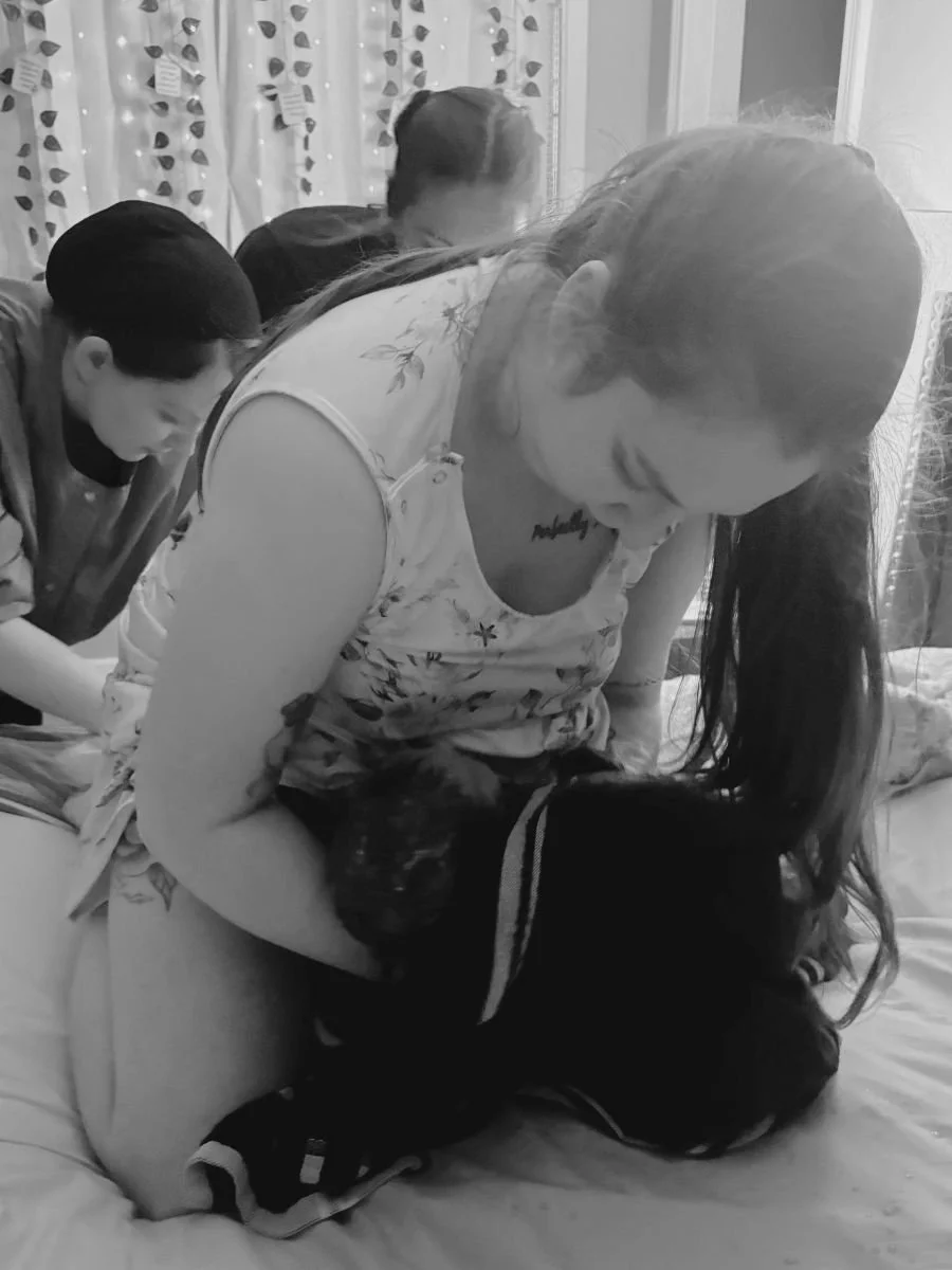 Doula, Hannah Servis, on her knees pulling her baby to her chest after having a homebirth with her midwife and sister standing behind and supporting her.
