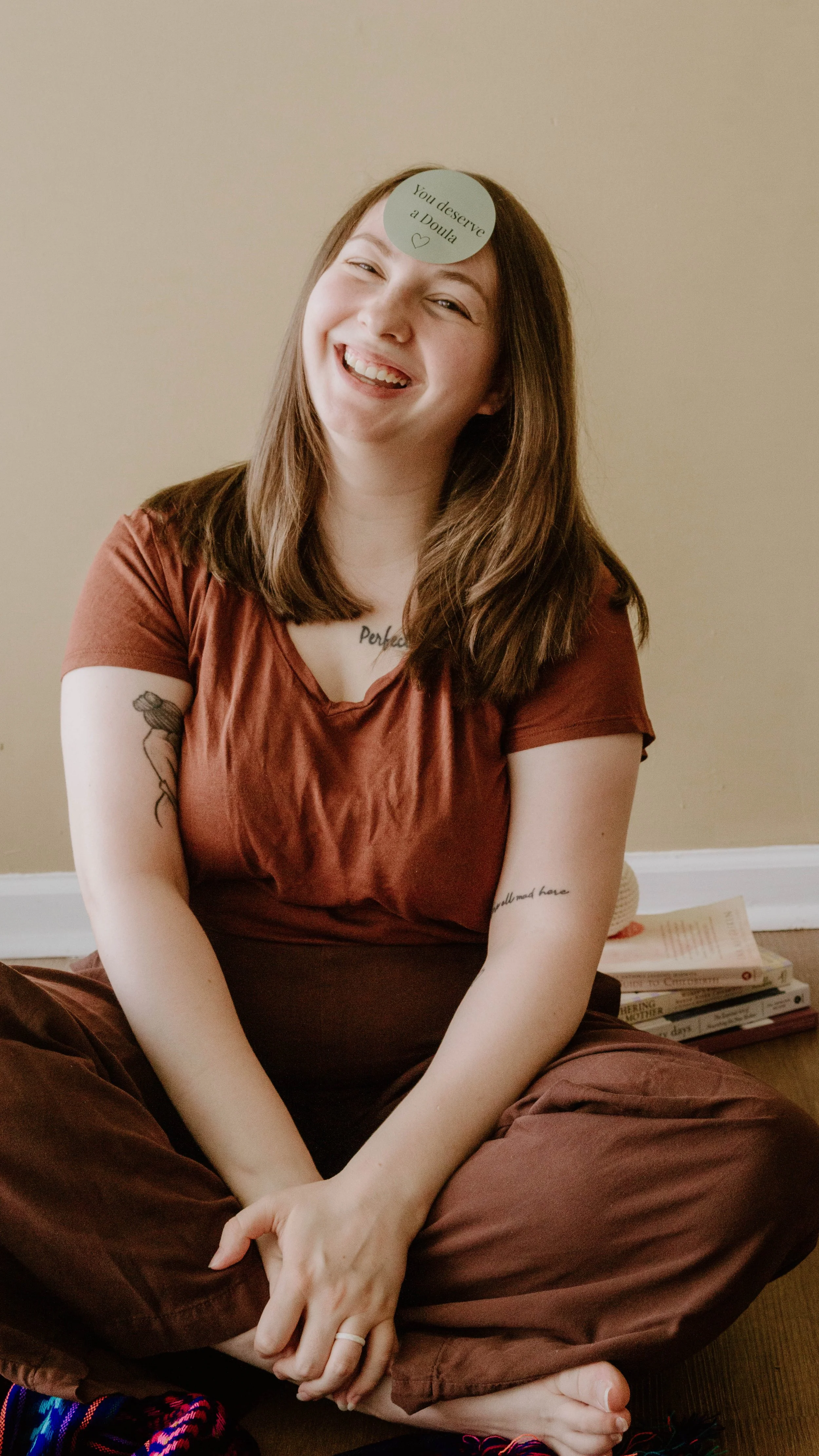 Birth & postpartum doula, Hannah Servis, sitting criss-cross on the floor laughing with a sticker on her forehead that reads "You deserve a doula".