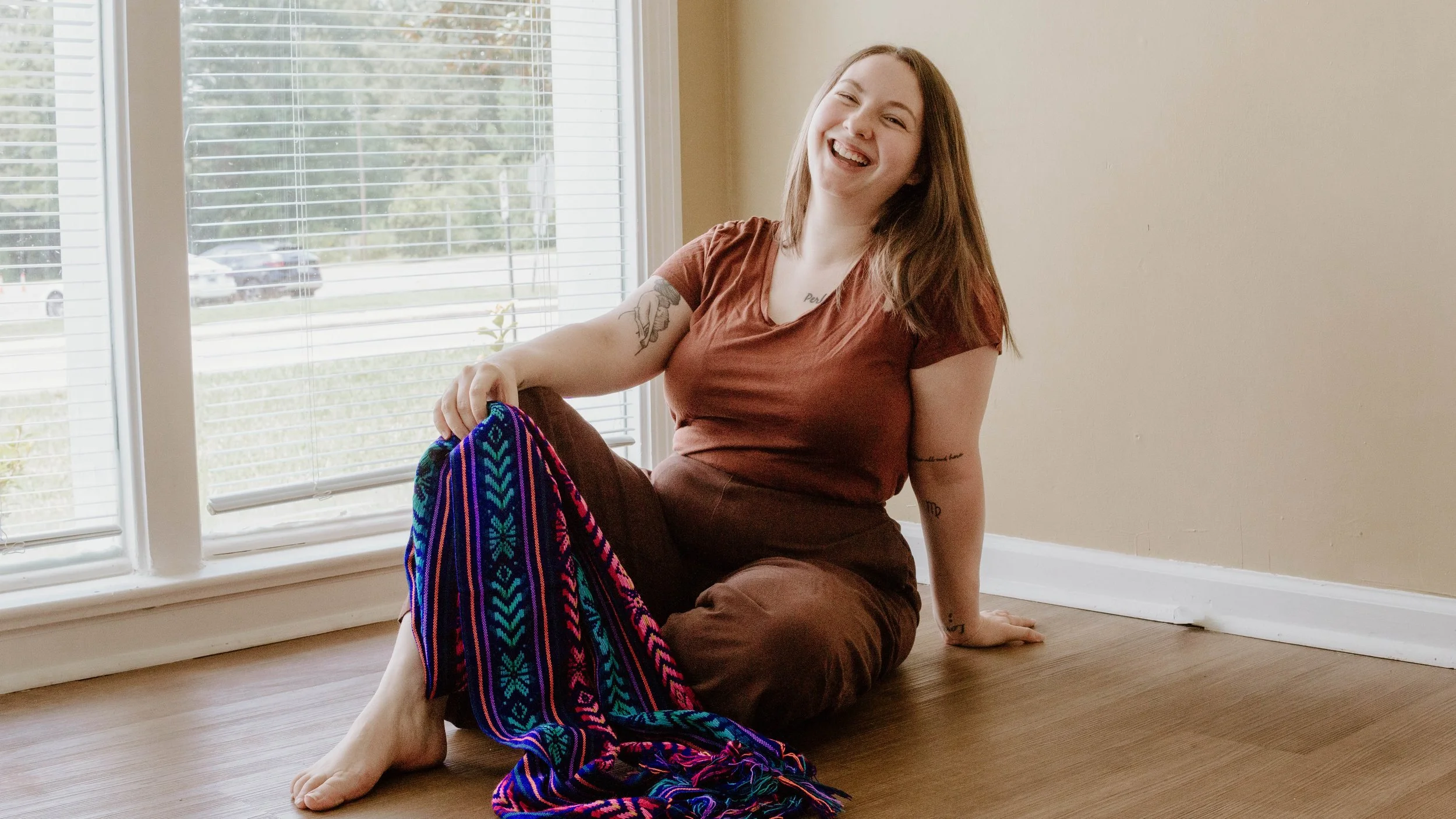 Birth Doula, Hannah Servis, sitting on the ground holding a rebozo in her hand with her head thrown back laughing as she thinks about all of the fun times she has had supporting births.