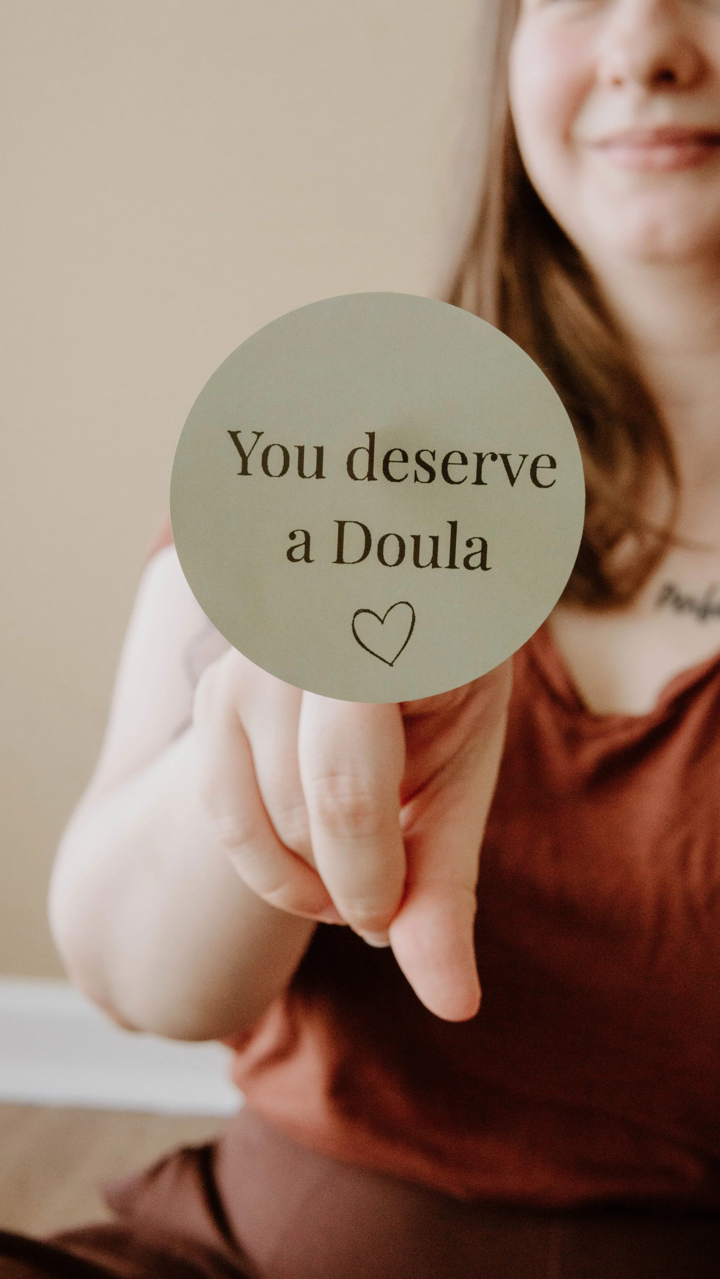 Birth and postpartum doula, Hannah Servis, holding a green circle sticker on her finger that says "You deserve a doula".