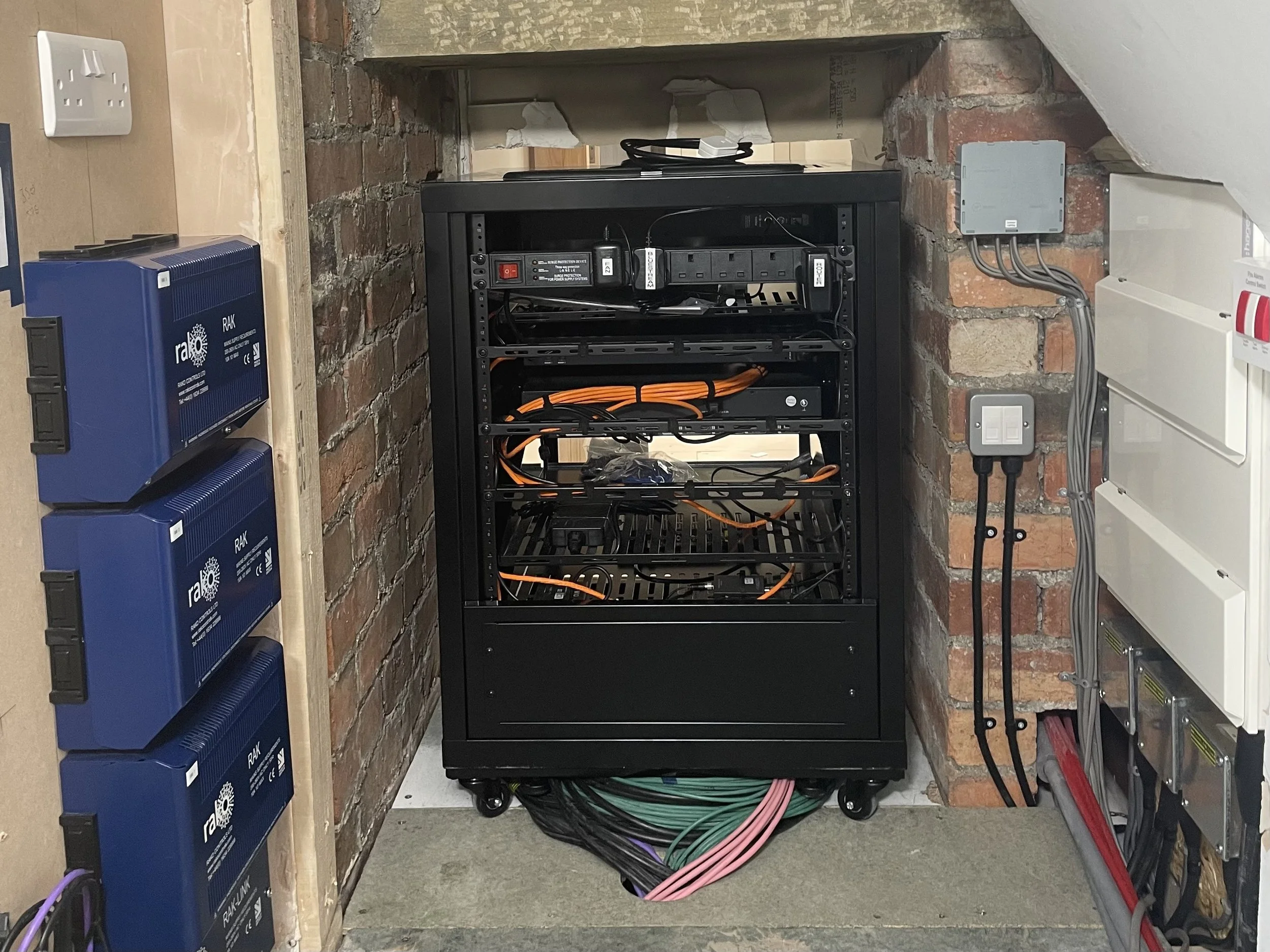 A server rack with network cables, power strips, and electrical connections, positioned against a brick wall with electrical outlets and circuit breakers nearby.