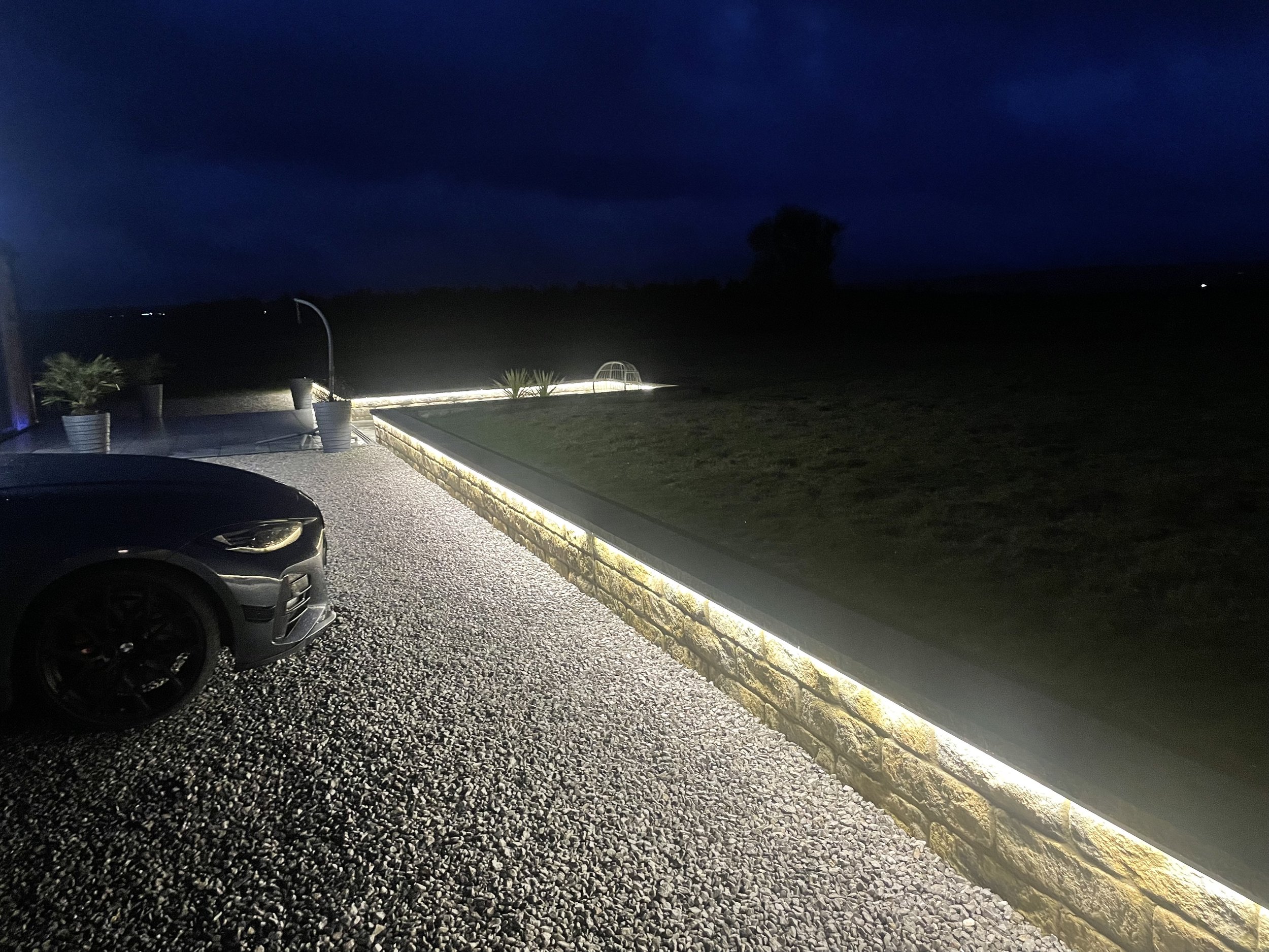 Nighttime outdoor scene with a dark sky, a tree in the distance, and a section of a driveway with a silver car, potted plants, and landscape parapet wall LED lighting.