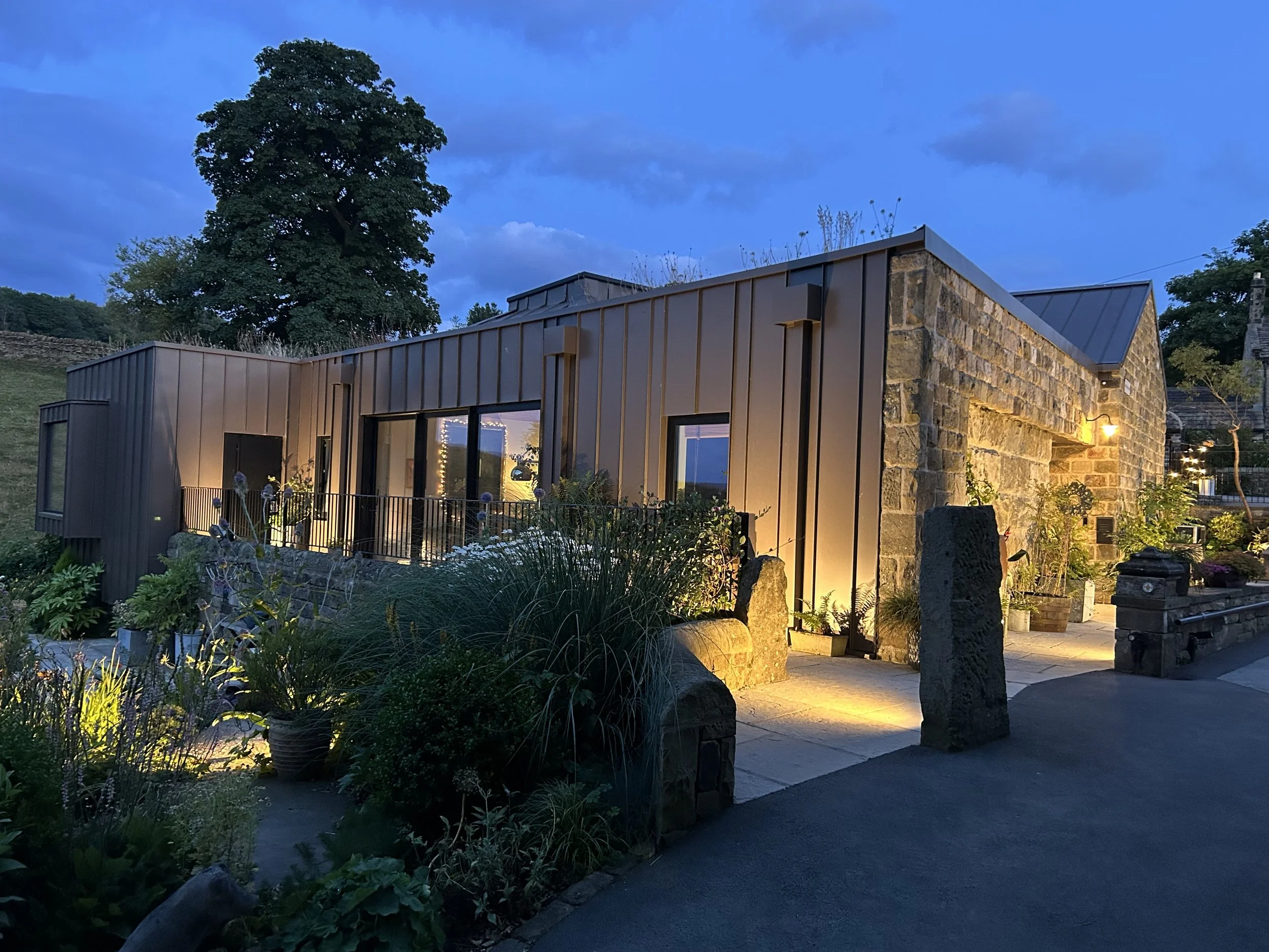 Exterior of a modern building with stone and zinc cladding, illuminated by outdoor lighting, with surrounding trees and a garden at dusk.
