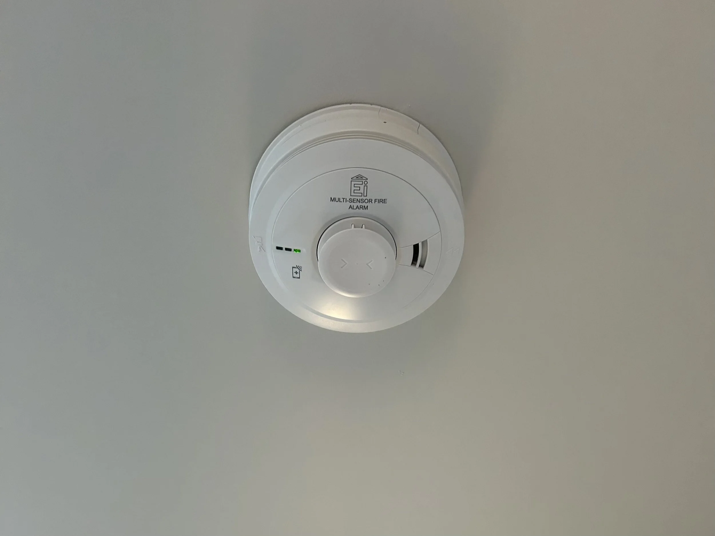 A white multi-sensor fire alarm mounted on a ceiling with a small green indicator light.