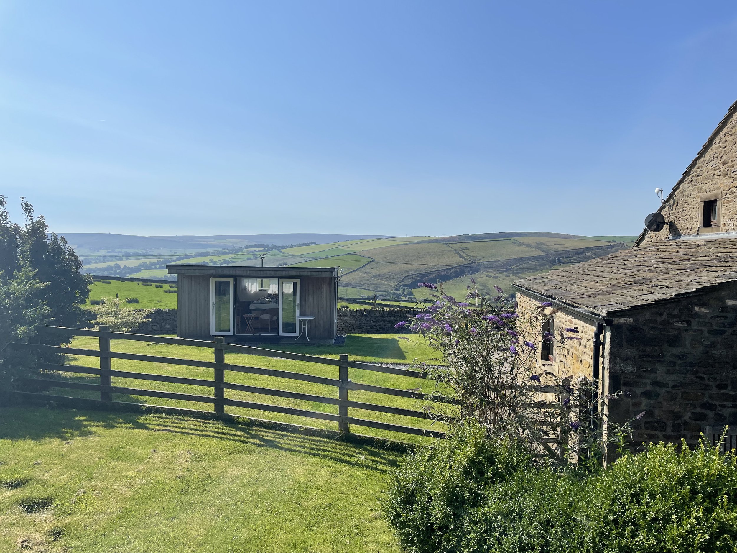 A scenic view of a rural landscape with rolling hills under a clear blue sky, a building with stone walls and a sloped roof on the right, and a small modern structure with sliding glass doors in the foreground, surrounded by greenery and a wooden fence.