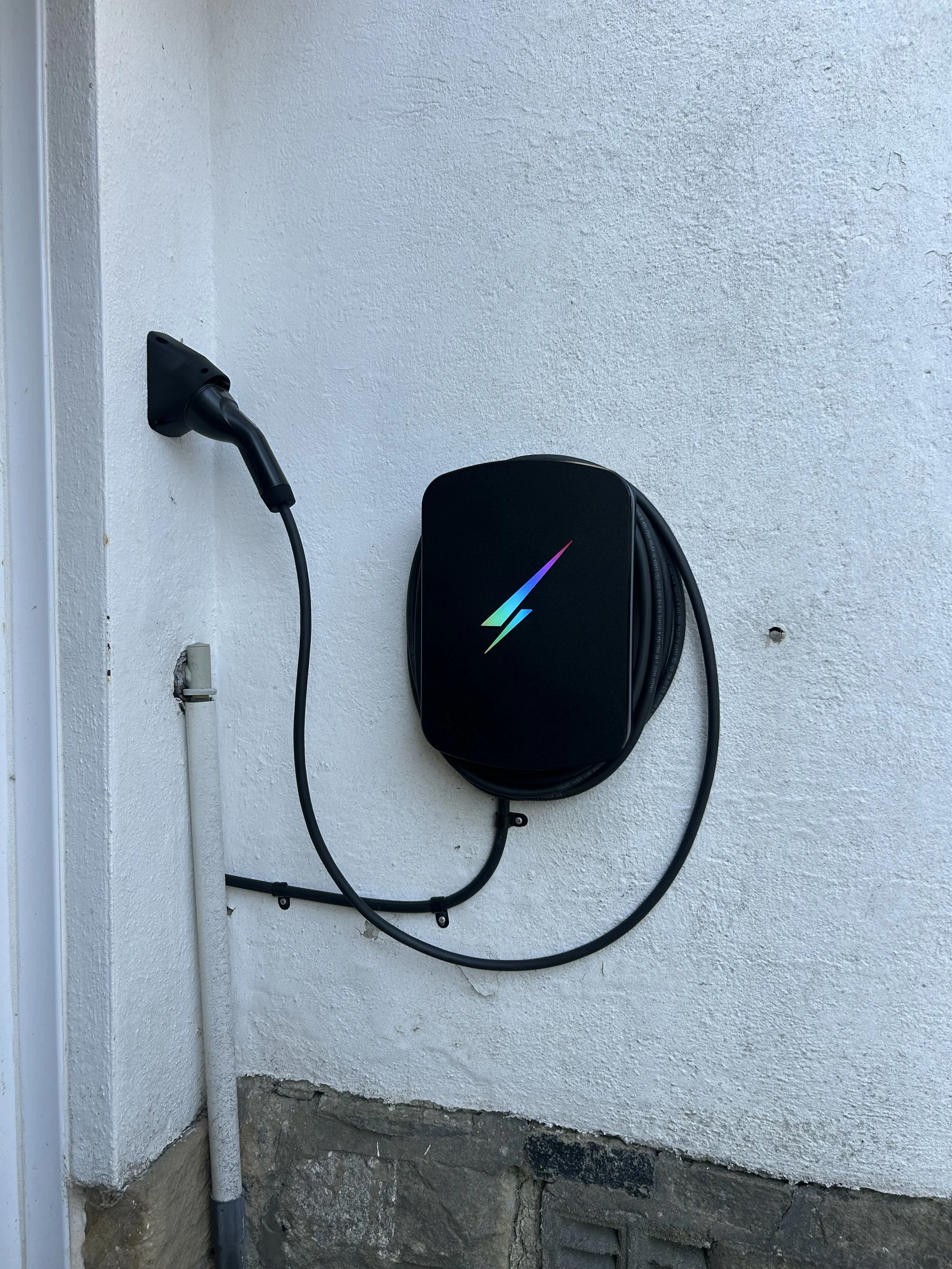 Electric vehicle charging station mounted on a white wall, with a black charging cable and plug connected, and a charging station case with illuminated logo.