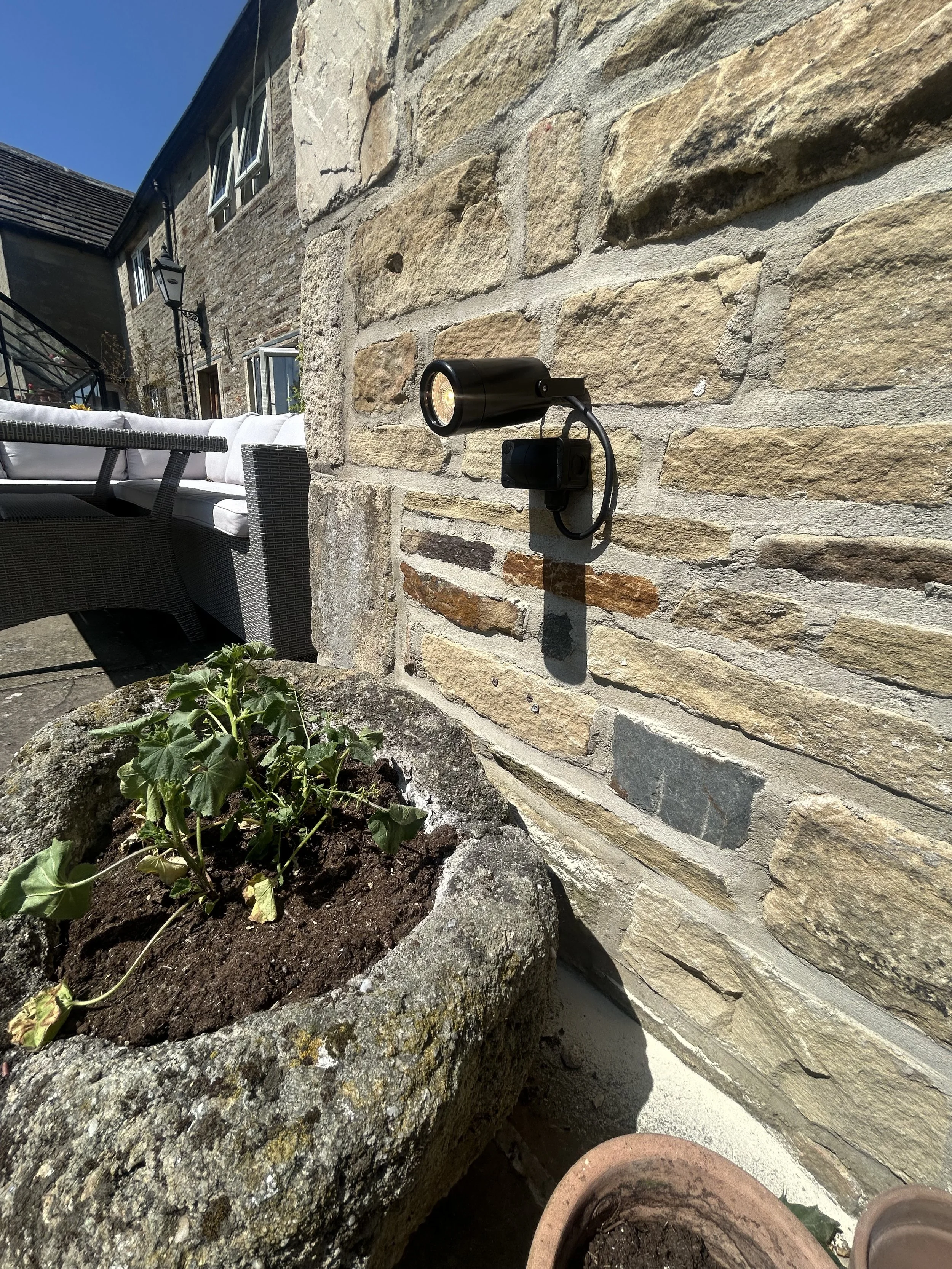 The image shows a stone wall with a black outdoor light fixture attached, casting a shadow, and a small garden bed with plants in pots and soil in front of the wall. There is a bench with white cushions and a pergola or outdoor structure in the background under a clear blue sky.